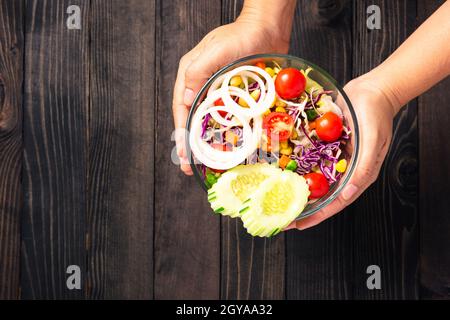 Draufsicht der Frauenhände, die die gesunde bunte Salatschüssel mit frischem Gemüse in einem Gericht auf schwarzem Holzhintergrund halten, Gesundheits-Snack f Stockfoto