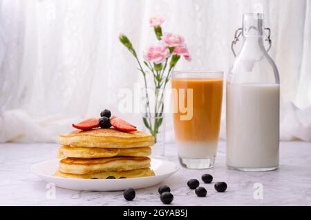 Stapel von Pfannkuchen mit frischen Erdbeeren und Heidelbeeren auf einem weißen Teller, einer Flasche Milch, einem Glas Milchtee und einer Vase mit rosa Blumen auf Marmor Stockfoto