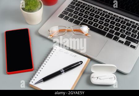 Geöffnetes Lehrbuch mit handgeschriebener TO-DO-LISTE in der Nähe von Laptop, schnurlosen Kopfhörern und Mobiltelefonen auf einem grauen Schreibtisch aus nächster Nähe Stockfoto