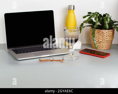 Intelligenter Arbeitsplatz - Laptop, isolierte Flasche, Pflanze, Handy, Gläser und ein Glas Wein auf grauem Schreibtisch aus nächster Nähe Stockfoto