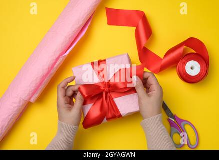 Weibliche Hände binden ein Seidenband auf einer umschlossenen Schachtel aus rosa Papier. Gelber Hintergrund, Draufsicht Stockfoto