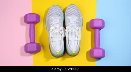 Ein Paar weiße Damen-Sneaker und lila Hanteln auf einem bunten Hintergrund, Sport Stockfoto