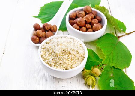 Mehl und Haselnüsse in zwei Schüsseln, ein Löffel mit geschälten Walnusskernen und ein Zweig aus Filbert mit Blättern auf dem Hintergrund von hellem Holzbrett Stockfoto