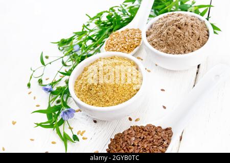 Leinenkleie und Mehl in zwei weißen Schalen, Samen in Löffeln, Leinenblätter und Blumen auf dem Hintergrund eines hellen Holzbretts Stockfoto