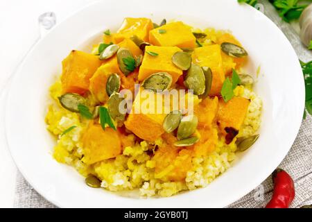 Hirsebrei mit würziger Kürbissauce und Samen auf einem Teller auf einer Serviette, scharfer Pfeffer, Ingwerwurzel, Knoblauch und Petersilie auf weißem Holzbrett Stockfoto