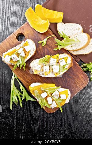 Bruschetta mit gebackenem Kürbis, gesalzenem Feta-Käse, Ricotta, Rucola und Gewürzen, Handtuch- und Gemüsescheiben auf Holzbrett-Hintergrund von oben Stockfoto