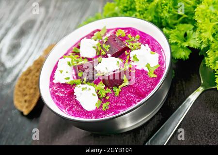 Suppenpüree aus Rote Beete, Kartoffel und Rahm mit gesalzenem Frischkäse in einer Schüssel auf Serviette, Brot und Petersilie auf schwarzem Holzbrett Hintergrund Stockfoto