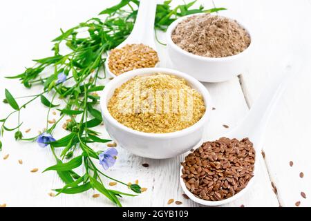 Leinenkleie und Mehl in zwei Schüsseln, Samen in Löffeln, Leinenblätter und Blumen auf dem Hintergrund eines weißen Holzbretts Stockfoto