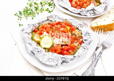 Rosafarbener Lachs mit Zucchini, Tomaten, Zwiebeln, Knoblauch und Thymian, in Folie auf einem Teller gebacken, Serviette, Gabel und Brot auf einem Holzbrett Hintergrund Stockfoto