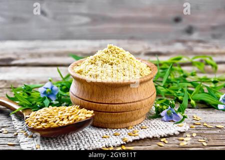 Leinenkleie in einer Schüssel, Samen im Löffel auf dem Sacken, Leinenblätter und Blumen auf dem Hintergrund eines alten Holzbretts Stockfoto