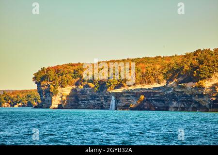 Pictured Rocks mit entferntem Wasserfall, der in den See fließt, mit Fallbäumen und rauschigen Wellen Stockfoto