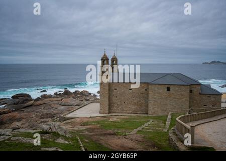 Wallfahrtskirche Virxe da Barca in Galicien mit Leuchtturm Cabo Vilan im Hintergrund in Galicien, Spanien Stockfoto