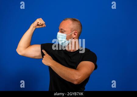 Junger gutaussehender Mann mit medizinischer Maske und schwarzem T-Shirt auf blauem Hintergrund isoliert. Starke Person zeigt Arm Muskel, selbstbewusst und stolz auf Macht zu Stockfoto