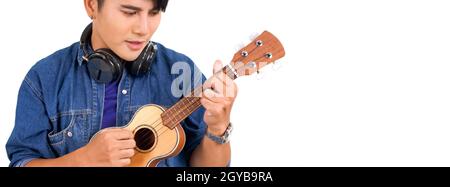 Junger asiatischer Mann mit Kopfhörern, der eine Ukulele-Gitarre spielt. Porträt auf weißem Hintergrund mit Studiolicht. Nahaufnahme Stockfoto