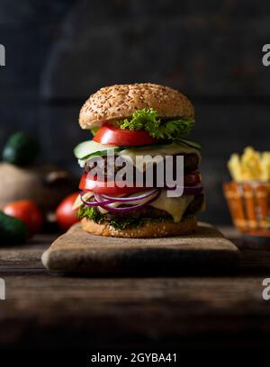 Hamburger mit Tomaten, Zwiebeln, Gurke, Salat und Schmelzkäse auf einem rustikalen Holzbrett mit pommes Frites und Gemüse im Hintergrund serviert Stockfoto