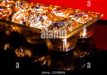 Eine Schachtel mit runden Bonbons in goldener Folie auf schwarzem und rotem Hintergrund. Verpackte Pralinen. Runder Kuchen. Süßes Essen. Süßes Geschenk. Goldene Farbe. Food-Foto. Stockfoto