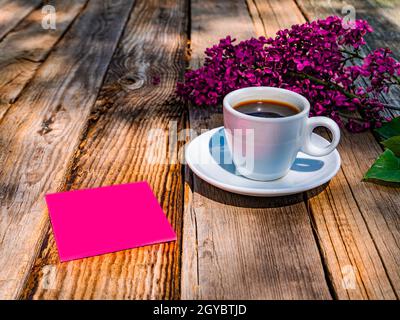 Eine Tasse Kaffee und ein Blatt Papier für Notizen neben einem Zweig Flieder auf einem Holztisch. Kaffeegetränk. Fliederbaumblüten. Tasse und Untertasse. Holztisch Stockfoto