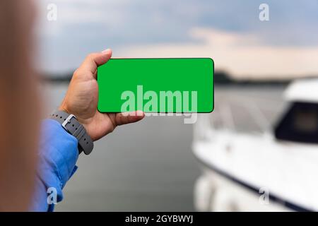 Mann hält Smartphone mit grünem Bildschirm in der Hand neben dem Fluss. Platz kopieren, Text, Foto oder Logo hinzufügen Stockfoto