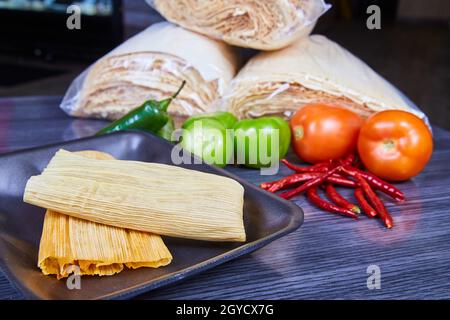 Tamales auf dem Tablett mit mexikanischen Zutaten aus Maisschalen, Tomaten und Paprika im Hintergrund Stockfoto