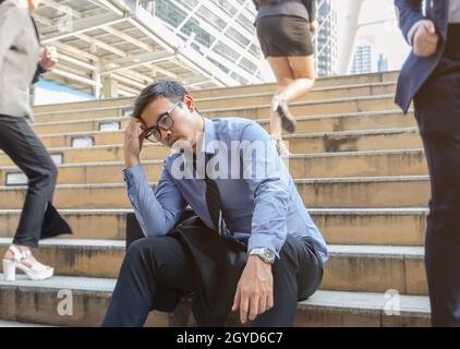 Deprimierter Geschäftsmann, der noch auf einer belebten Treppe im Freien sitzt, während verschwommene Geschäftsfrauen in der Stadt an ihm vorbeigehen. Arbeitsloser Mann verzweifelt wirtschaftliche Kris Stockfoto