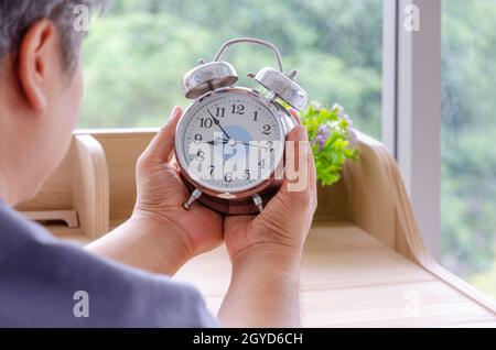 Älterer Mann, der am Schreibtisch sitzt und einen weißen Wecker hält Stockfoto