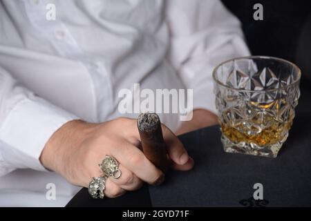 Eleganter Mann in weißem Hemd hält eine rauchenden kubanischen Zigarre. Ein Glas Whiskey auf dem Tisch Stockfoto