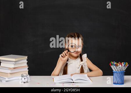 Nettes Schulmädchen suchen durch Vergrößerungsglas im Klassenzimmer auf Blackboard Hintergrund Stockfoto
