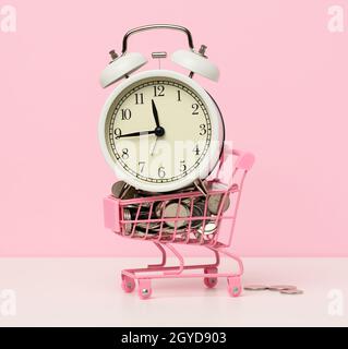 Rund-um-die-Uhr-Wecker in einem Mini-Einkaufswagen mit Wechsel auf einem weißen Tisch. Konzeptzeit ist Geld, Geldverschwendung und Armut Stockfoto