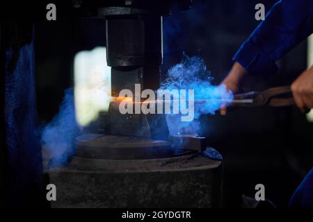 Kaukasischer Handwerker in Uniform, der in der Werkstatt erhitztes Metall unter die Pressenmaschine setzt. Nahaufnahme des Arbeitsprozesses bei FORGE. Stockfoto