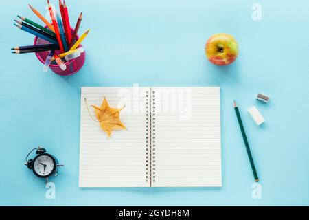 Leerer Notizblock und Schulbedarf, Wecker, Apfel und gelbes Herbstblatt auf blauem Hintergrund. Zurück zum Schulkonzept. Draufsicht, flaches Lay, Mock Up. Stockfoto