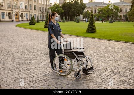In voller Länge Aufnahme einer fürsorglichen jungen Krankenschwester mit Schutzmaske während der Covid19-Pandemie, die im Park in der Nähe der Klinik mit einem Rollstuhl im Freien stand Stockfoto