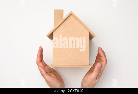 Weibliche Hände gefaltet zueinander über einem hölzernen Miniatur-Modellhaus auf weißem Hintergrund. Immobilienversicherungskonzept, Umweltschutz, Stockfoto