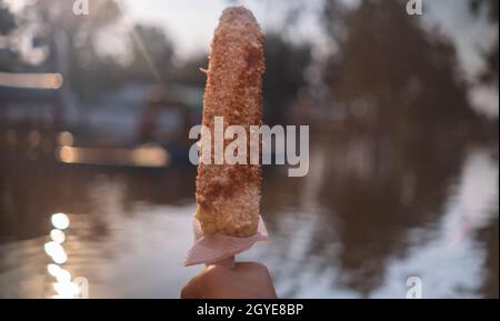 Hand hält traditionellen Mais mit Mayonnaise und Chilipulver über dem See Stockfoto