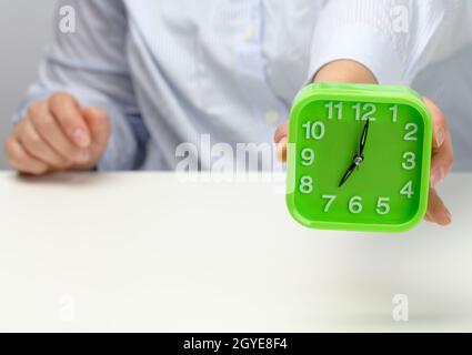 Die Hand der Frau hält einen grünen quadratischen Wecker, die Uhr zeigt sieben Uhr morgens an. Wachen Sie früh auf, und beginnen Sie den Tag Stockfoto