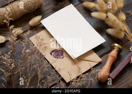 Leere Papierkarte auf versiegeltem Umschlag und Holztisch mit getrockneten Pflanzen, Nahaufnahme. Boho-Mock-up-Szene mit Grußkartenvorlage Stockfoto