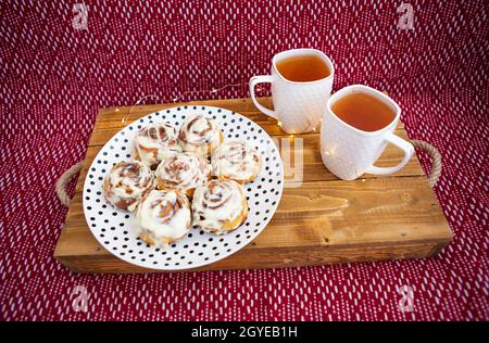 Zwei Tassen schwarzen Tee stehen auf einem Holztablett. Frische und duftende Zimtrollen liegen ganz nah auf einem Teller mit Tupfen, schöner Morgen. Nahaufnahme. Stockfoto