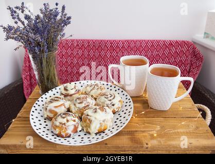 Zwei Tassen schwarzen Tee stehen auf einem Holztablett, ein Bouquet von getrocknetem Lavendel. Frisch und duftend Zimtrollen aus nächster Nähe liegen auf einem Teller mit Tupfen, Stockfoto