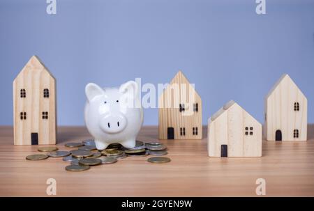 Ein weißes Sparschwein wird auf einen Haufen Münzen gelegt. Und daneben ist ein hölzernes Modellhaus auf weißem Hintergrund. Die alle auf einem Holztisch stehen Stockfoto