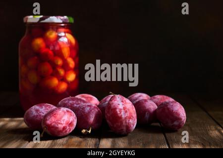 Frische Pflaumen und ein Glas Pflaumenkompott auf einem dunklen Holztisch, selektiver Fokus auf die zentrale Pflaume, geringe Schärfentiefe Stockfoto