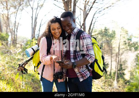 Glückliches, vielfältiges Paar mit Rucksäcken, das Smartphone beim Wandern auf dem Land benutzt Stockfoto
