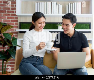 Asiatische Frau in weißem Langarm-T-Shirt gab ihrem Freund eine Tasse Kaffee, während der Mann einen Laptop in der Hand hielt. Junge Liebhaber verbringen Zeit miteinander Stockfoto