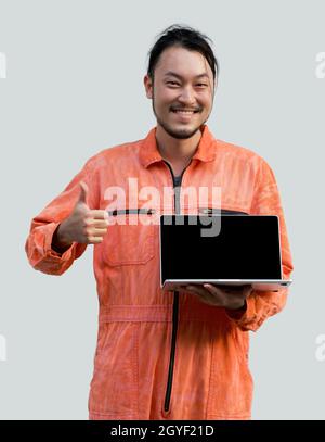 Der Chefmechaniker in einer orangefarbenen Uniform, der einen Laptop mit leerem Bildschirm hält. Mit den Daumen nach oben stehend. Porträt mit Studiolicht. Stockfoto