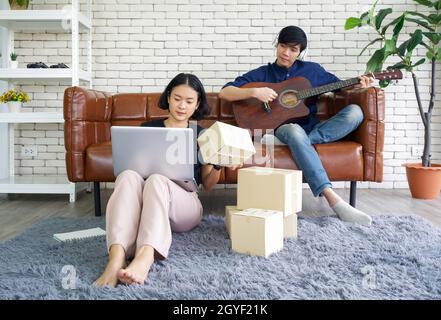 Junge Liebhaber verbringen Zeit zusammen auf Urlaub im Wohnzimmer. Asiatischer Mann mit Kopfhörern spielt eine akustische Gitarre, während das Mädchen das Paket überprüfen Stockfoto