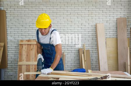 Der asiatische Zimmermann trägt einen gelben Harthut und schneidet in der Holzbearbeitungsanlage mit einer elektrischen Handsäge Sperrholz. Ein Schreibtisch mit Handwerkzeugen und Holzpfählen. Stockfoto