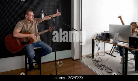 Der junge kaukasische Künstler mit Akustikgitarre überprüft die Korrektheit des Audiorecorders, bevor er mit dem Toningenieur beginnt. Musiker produzieren Stockfoto