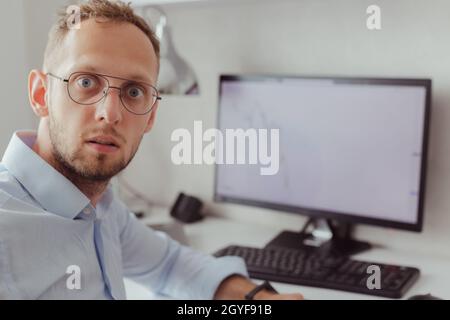 Porträt eines überraschten Mannes in Brille und Bürohemd auf dem Hintergrund des Monitors mit Grafik des fallenden Marktes. Börsenkrise. Kryptowährung. Lott Stockfoto