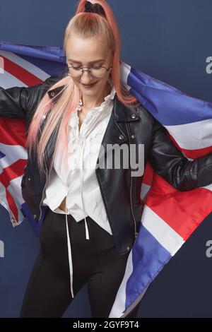 Junge Frau Expat in Brillen mit Großbritannien Flagge. Studioaufnahme. Hipster-Lehrerin in englischer Sprache. Fashionly gekleidete Frau mit rosa Haaren in bla Stockfoto