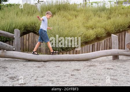 Der blonde Junge geht auf einem Holzbalken auf einem Spielplatz in einem öffentlichen Park. Stockfoto