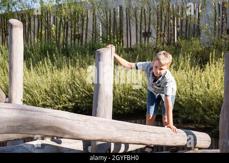 Der blonde Junge geht auf einem Holzbalken auf einem Spielplatz in einem öffentlichen Park. Stockfoto