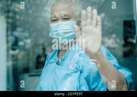 Asiatische alte ältere Patientin mit Maske, die während einer Covid19-Coronavirus-Pandemie aus dem Fenster schaut, um sich in Quarantäne zu begeben, bleibt zu Hause. Stockfoto